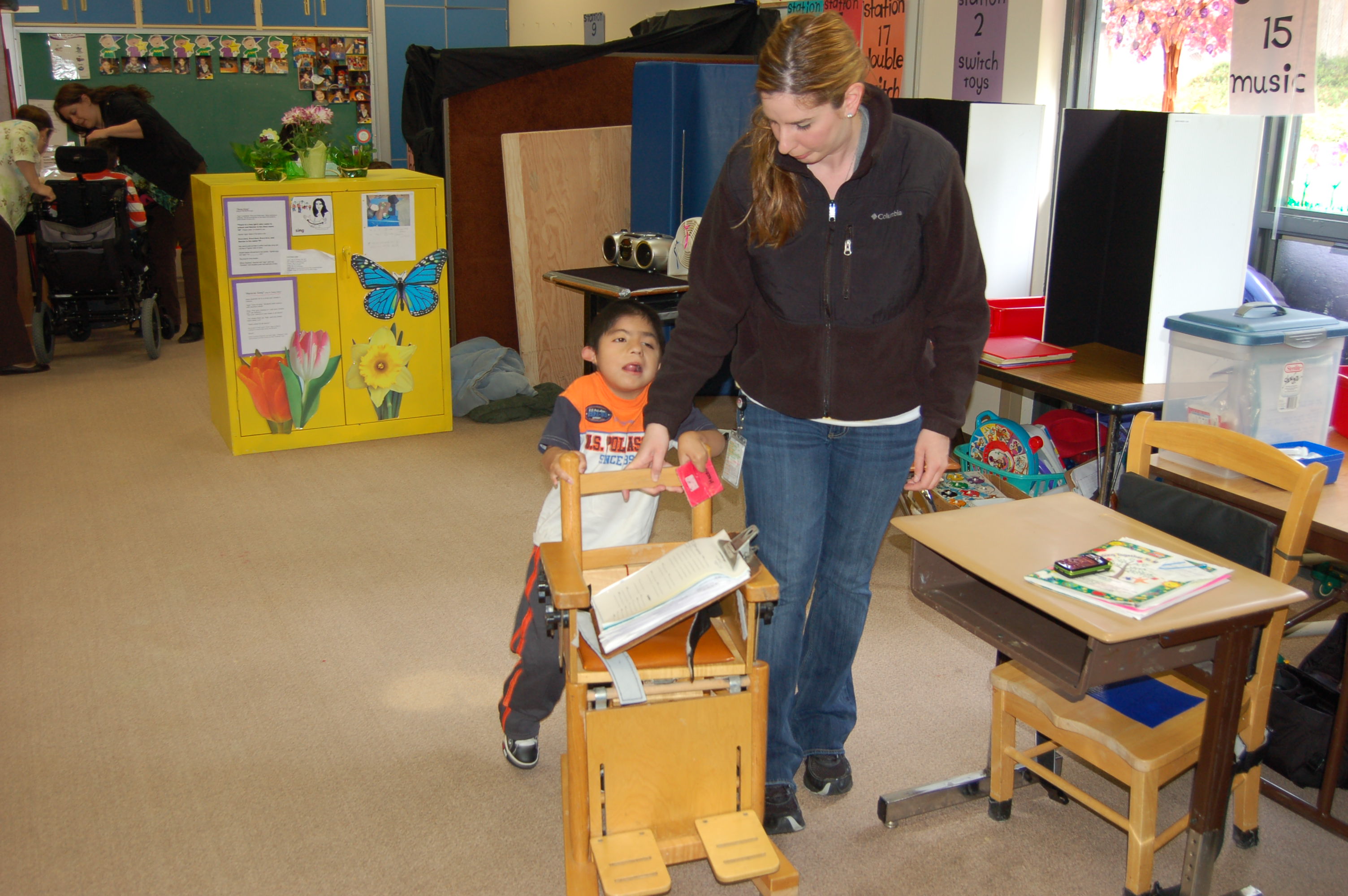 Un niño en un aula se toma de una silla especializada para apoyarse mientras camina. Le ayuda un adulto que también tiene la mano en el respaldo de la silla.