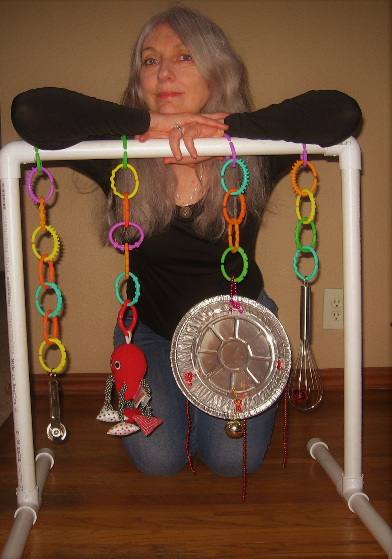 A woman is kneeling on a wood floor behind a homemade pvc pipe frame. She is leaning forward with her arms resting on the top of the frame. Hanging from it are four colored plastic chains. At the end of each chain is an object—a measuring spoon, a stuffed