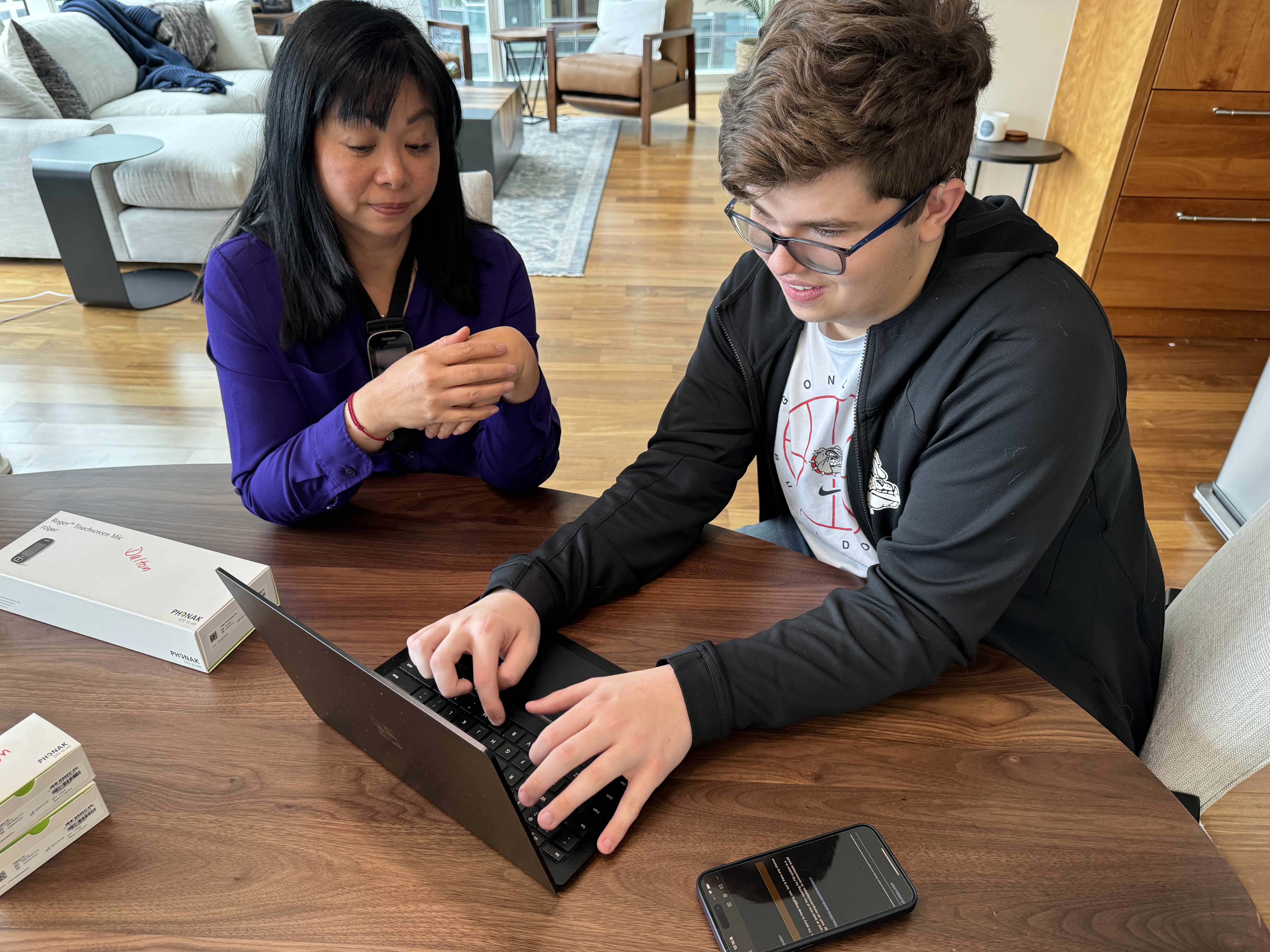 A young adult male who is deafblind sits in front of a laptop. He talks with an adult female seated beside him.