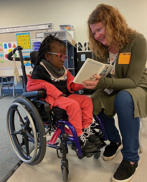 A young female student who is deaf-blind sits in a wheelchair. Her teacher sits beside her holding a braille book. The student reaches out to touch the book.