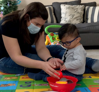 A toddler who is deafblind sits on a colorful floor matt with an adult female. Using her finger under his hand, they explore colorful objects in a bowl.