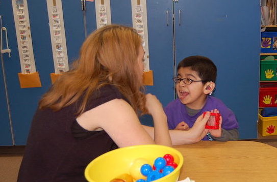 An aide and young boy are sitting at a table playing with a plastic toy.