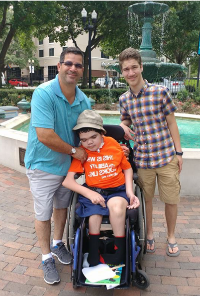 A young boy who is deafblind sits in his wheelchair in a park. His father and brother stand behind him. His father supports him by placing his hands on the boy’s arm and around his back.