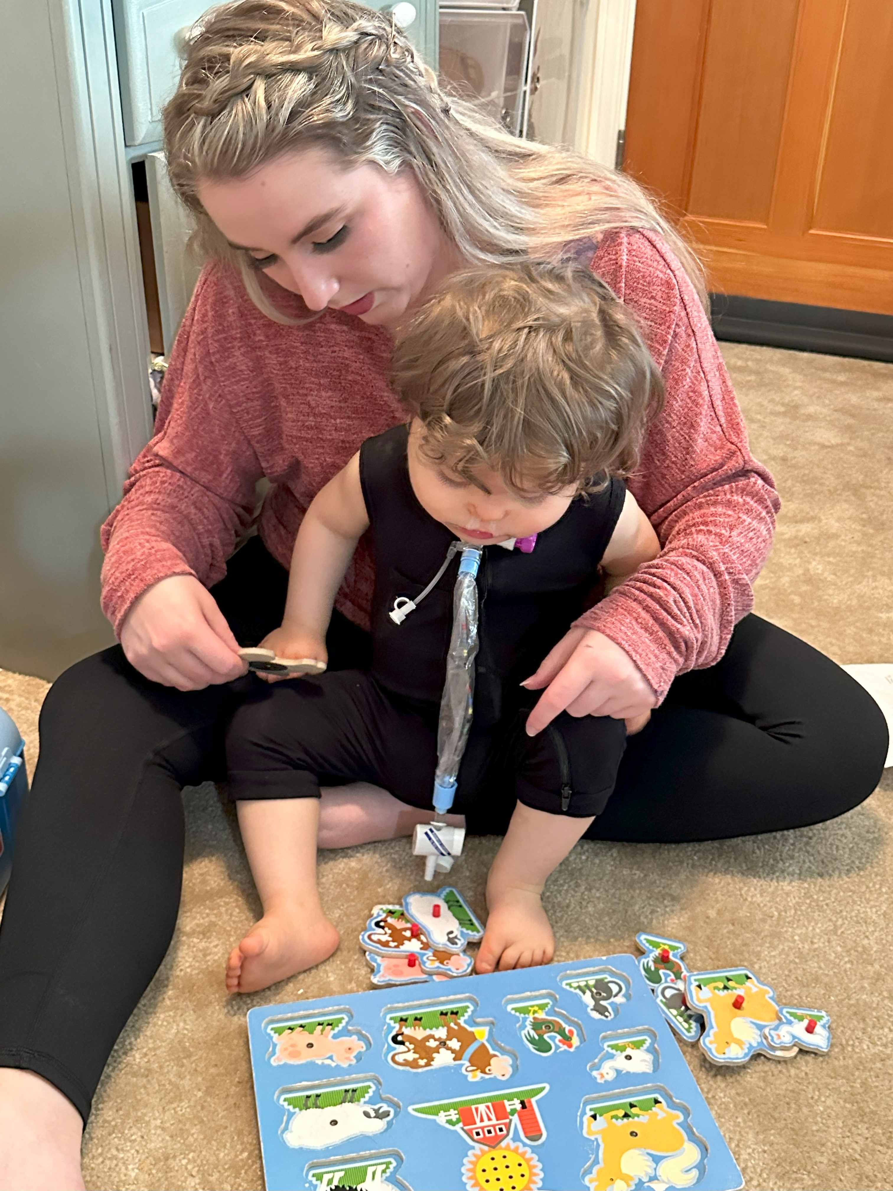 A young boy who is deafblind sits on the floor with his mom and plays with a puzzle. He has a tracheostomy tube.